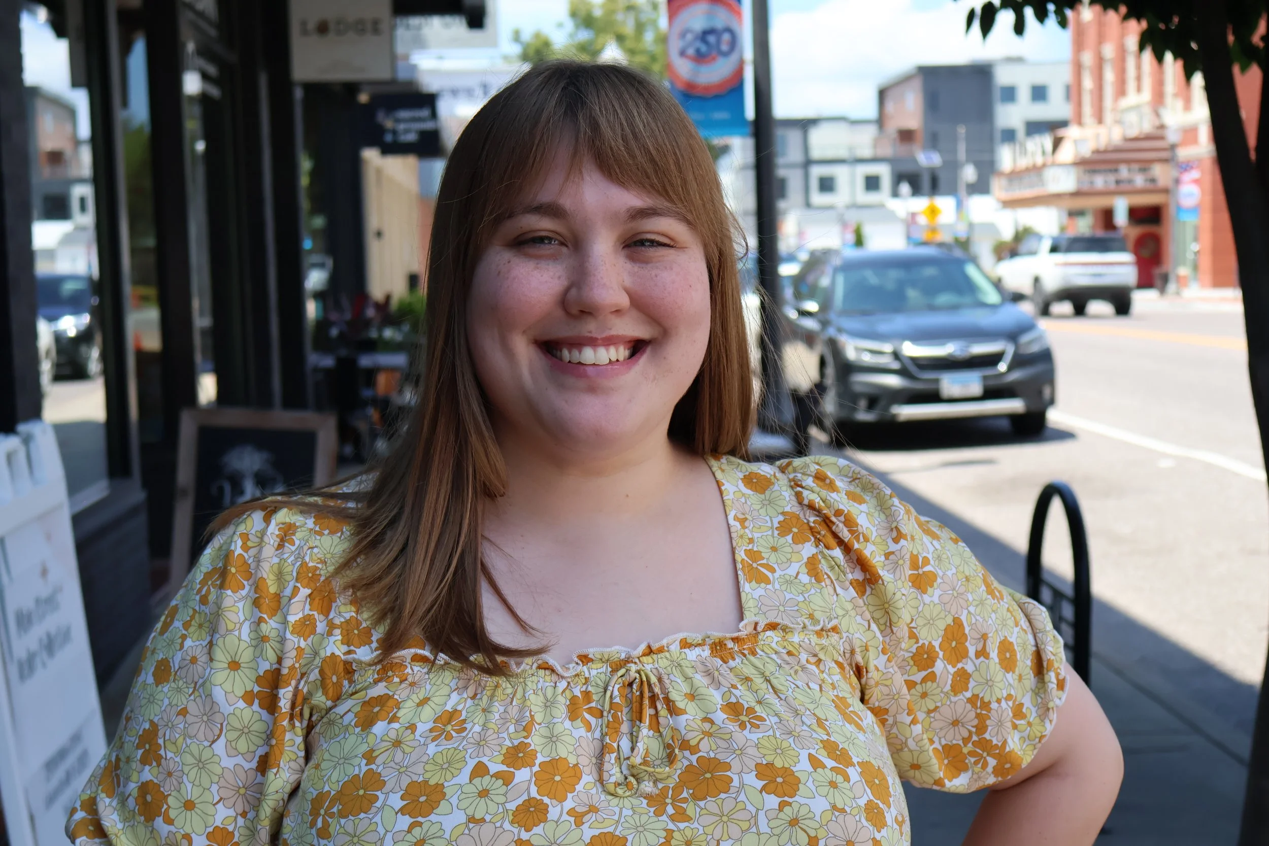 A young woman with red hair and freckles smiling outdoors, wearing a yellow floral dress. She is standing on a sidewalk in front of a shop, with parked cars and buildings in the background.