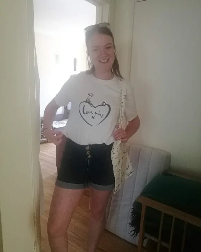 Young woman with long hair, wearing a white T-shirt with 'Love wins' inside a heart drawing, black shorts, smiling, standing in a doorway, holding a canvas tote bag, next to a bed or cushion in a room with wooden flooring.