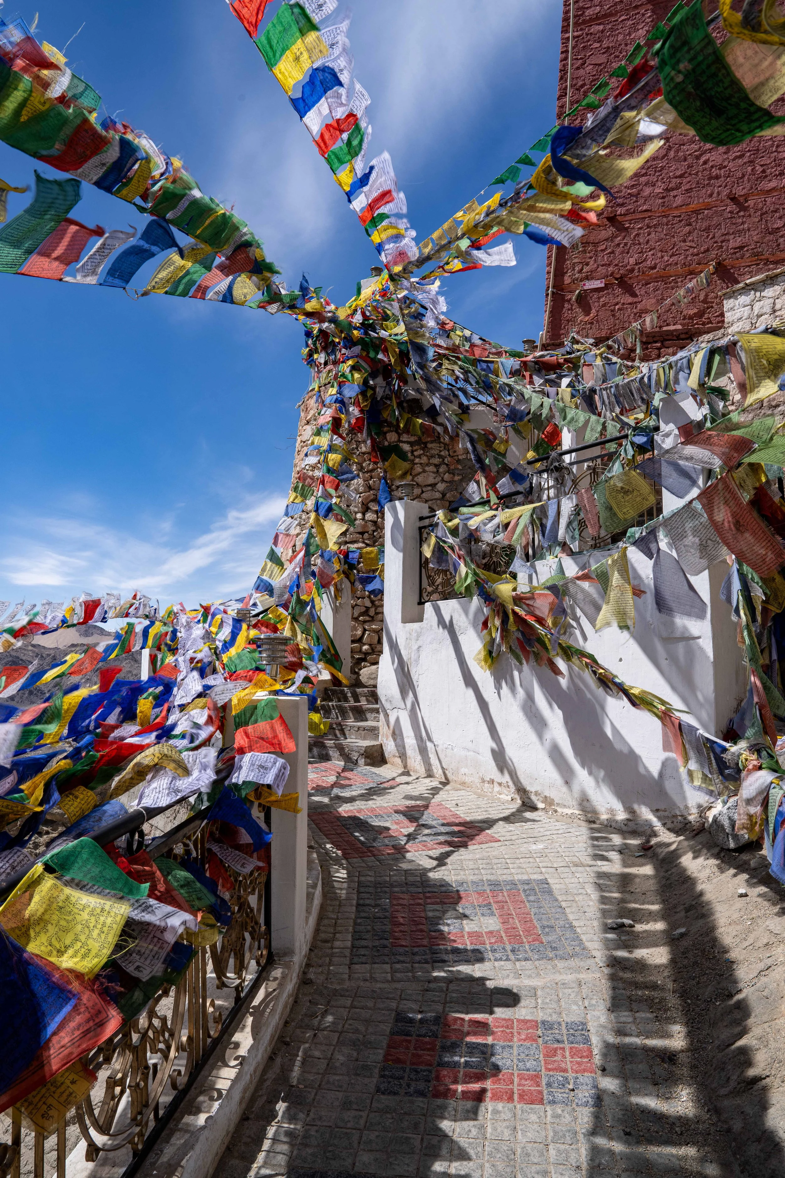 Colorful prayer flags hanging across a narrow street with shadows cast on the cobblestone pavement, a brick wall on the right, and a stone wall at the end of the street, under a bright blue sky.