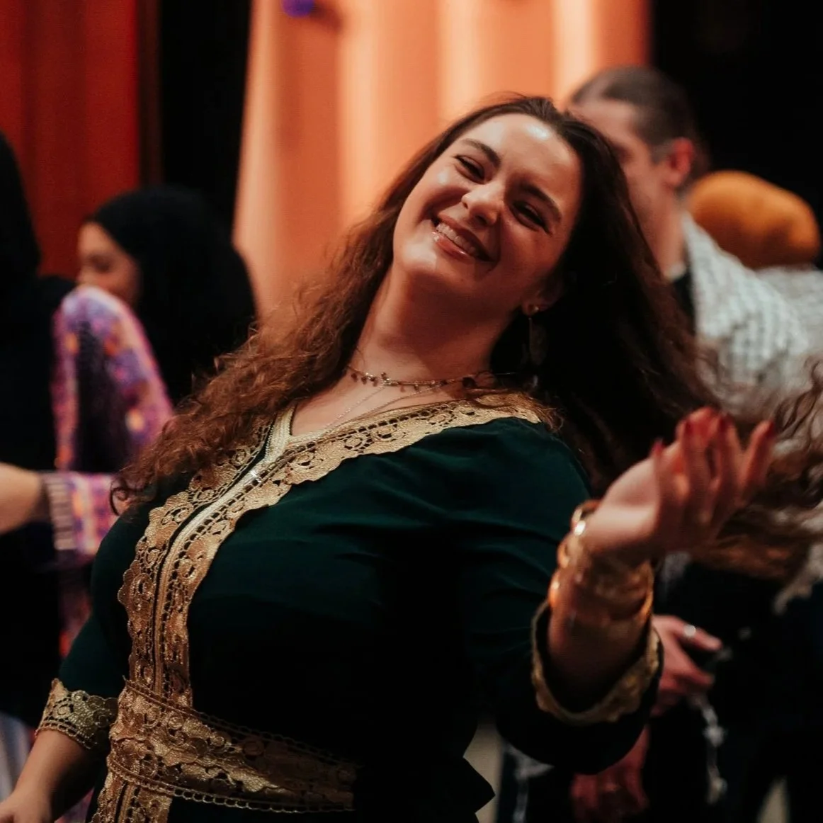 A woman smiling and dancing at a social event, wearing a black dress with gold embroidery, with people in the background.