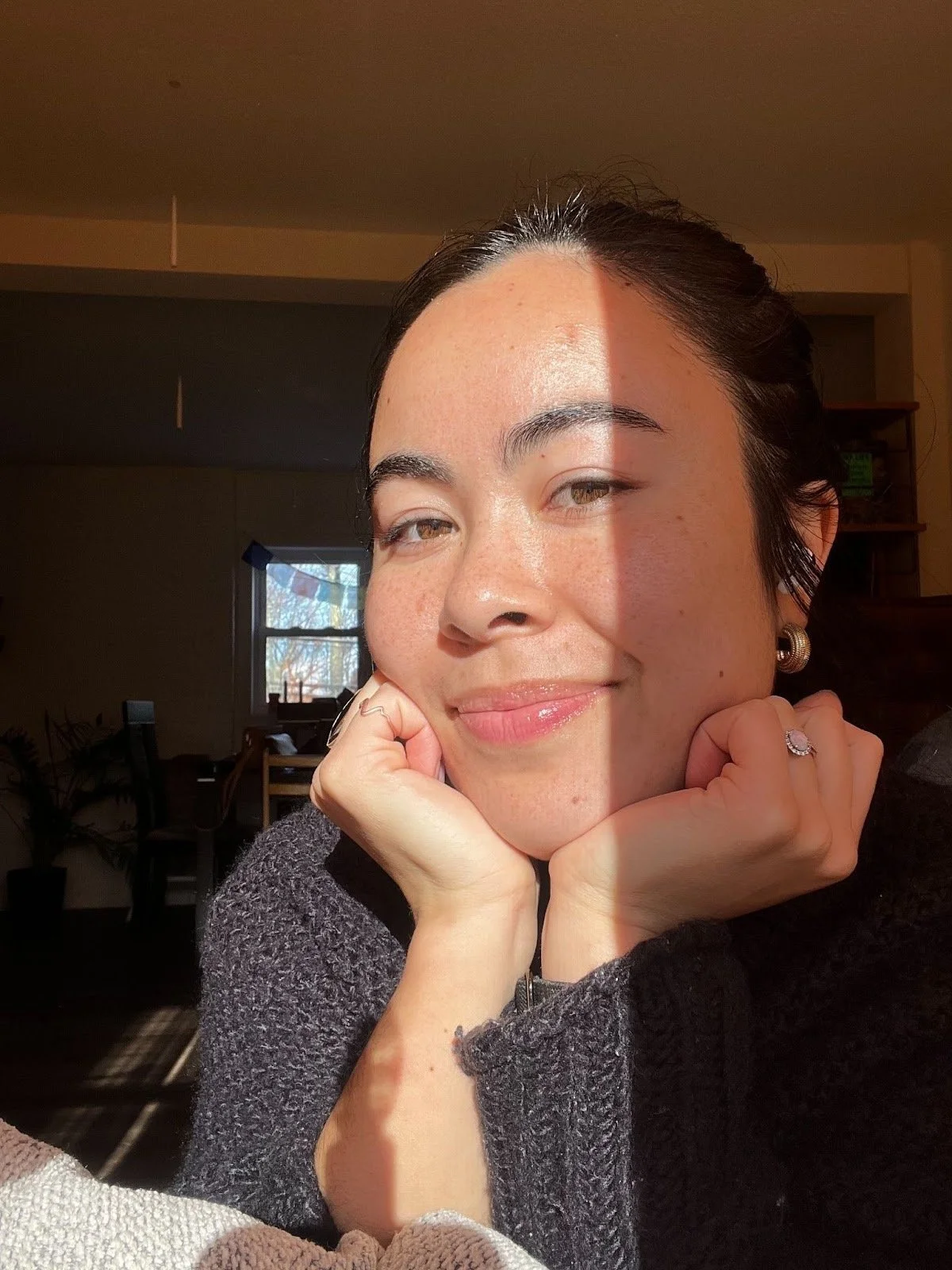 A woman with dark hair, light skin, and brown eyes, smiling gently, resting her chin on her hands, with sunlight illuminating her face indoors.