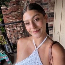 A young woman with dark hair in braids standing outdoors on a balcony, wearing a white sleeveless top and smiling.