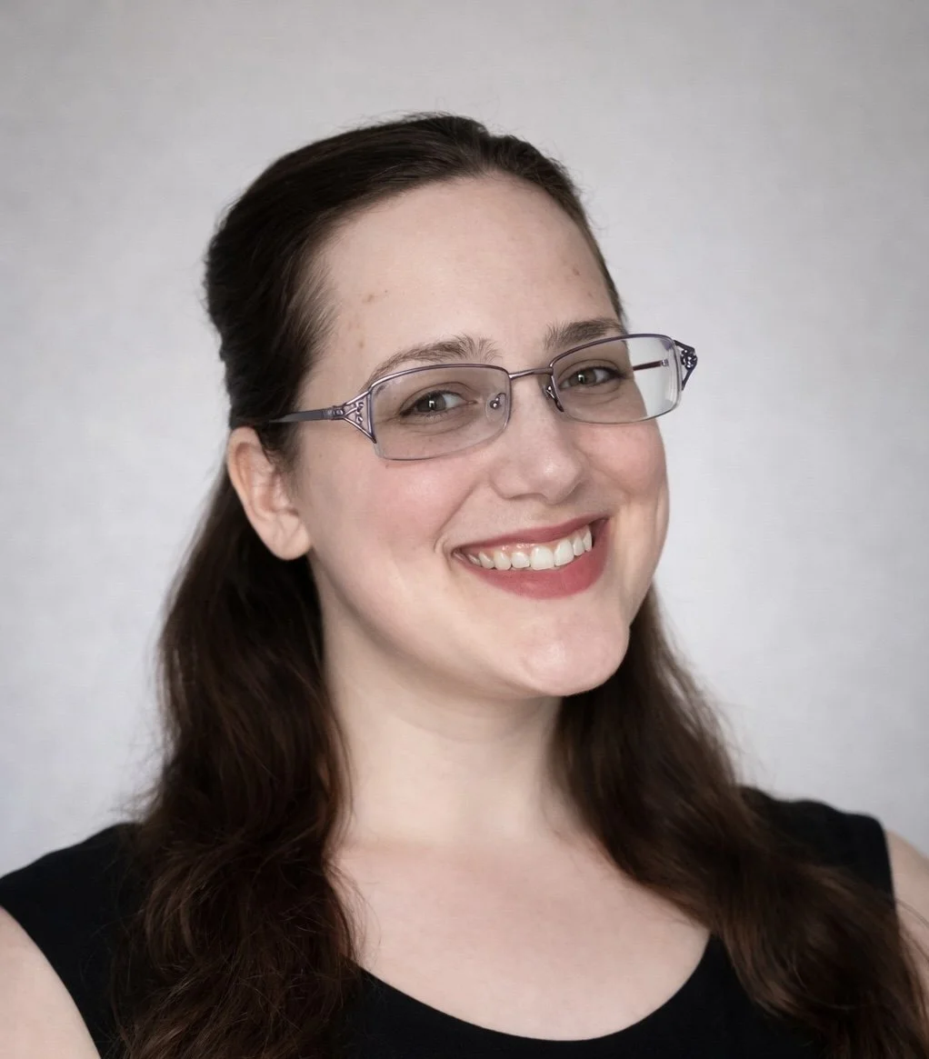 A woman with brown hair, glasses, and a black sleeveless top, smiling at the camera against a light gray background.