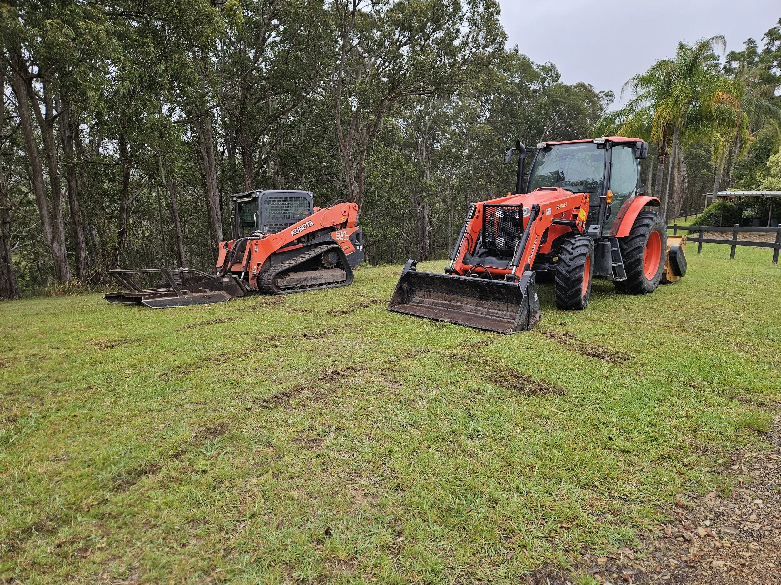 Two orange construction tractors on a grassy field near a wooded area with tall trees and cloudy sky.