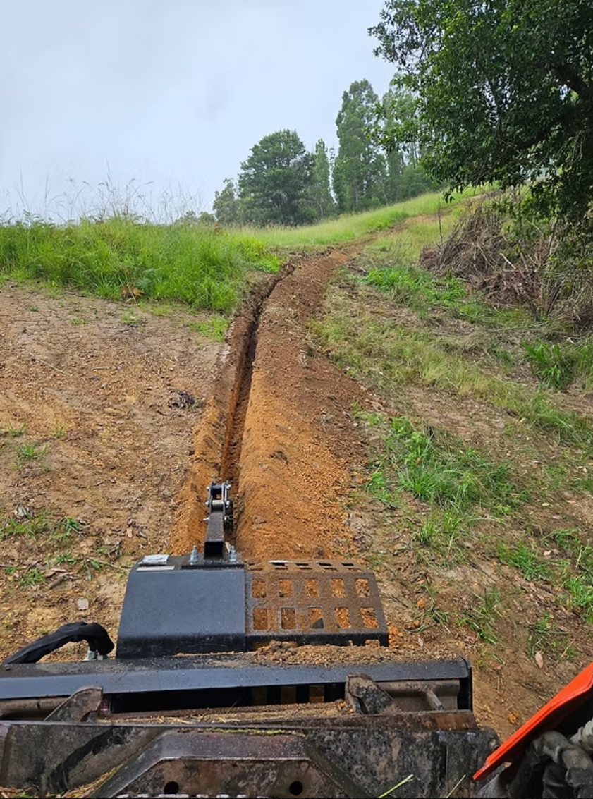 A machine excavating a narrow trench along a grassy hillside on a dirt path in a rural area with trees and cloudy sky in the background.
