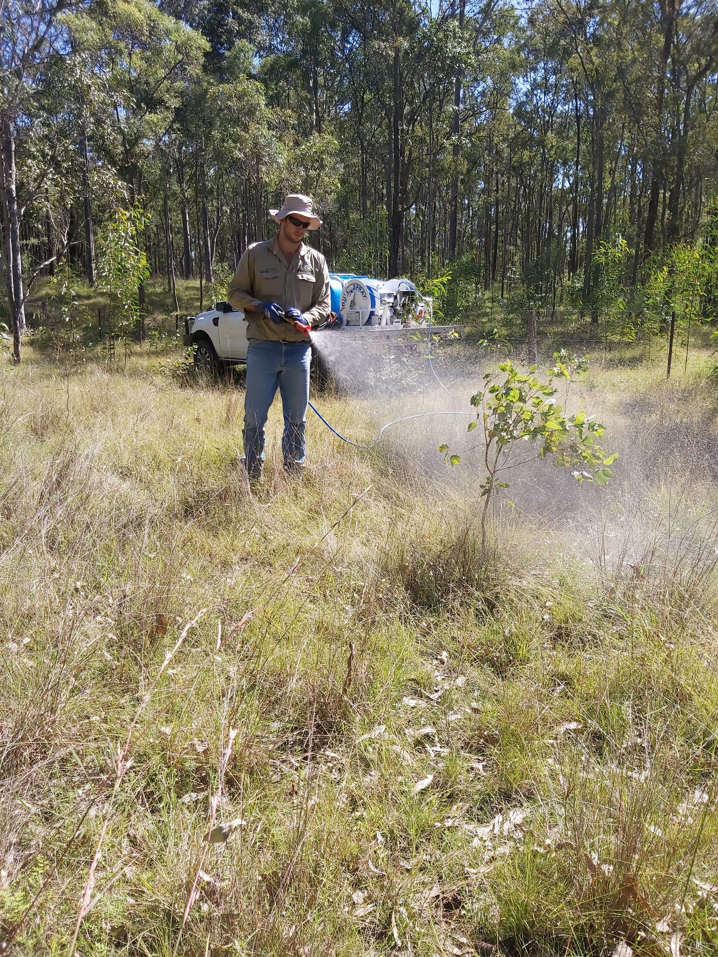 A person wearing a wide-brimmed hat, sunglasses, and gloves operating equipment in a grassy, wooded outdoor area.