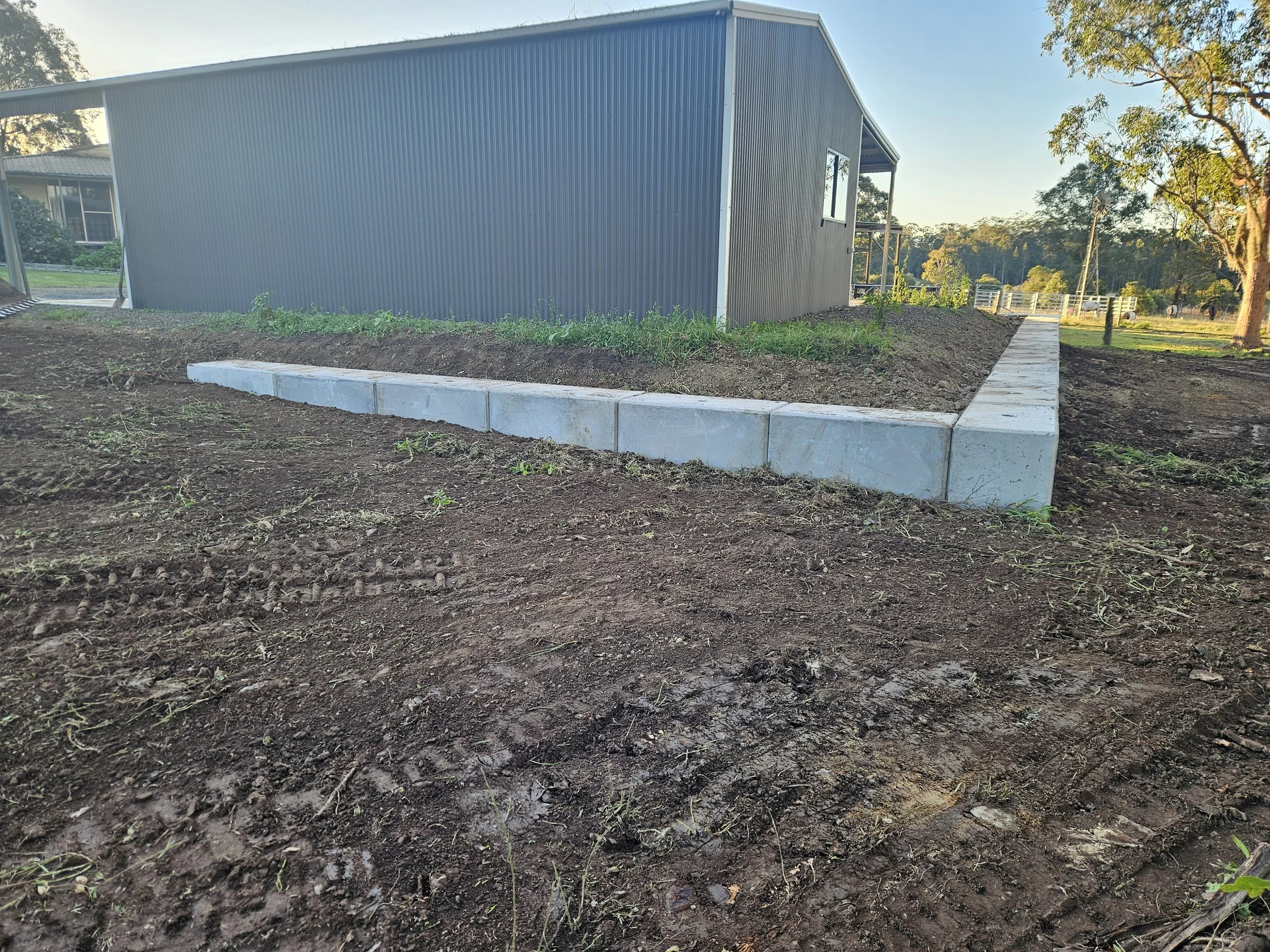 View of a gray metal building on a rural farm with a dirt and grass area in the foreground, trees in the background, and a clear sky.