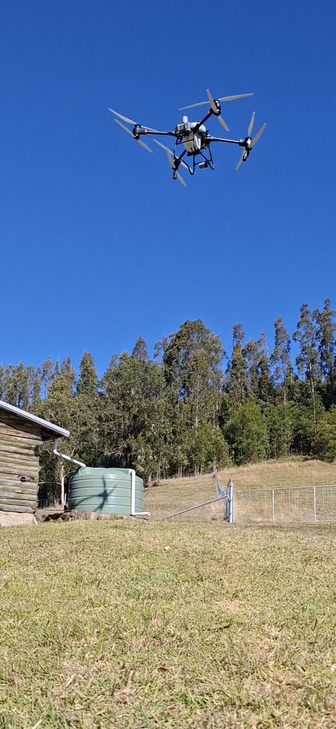 A drone flying in the sky above a rural area with trees, a water tank, and a small wooden structure.