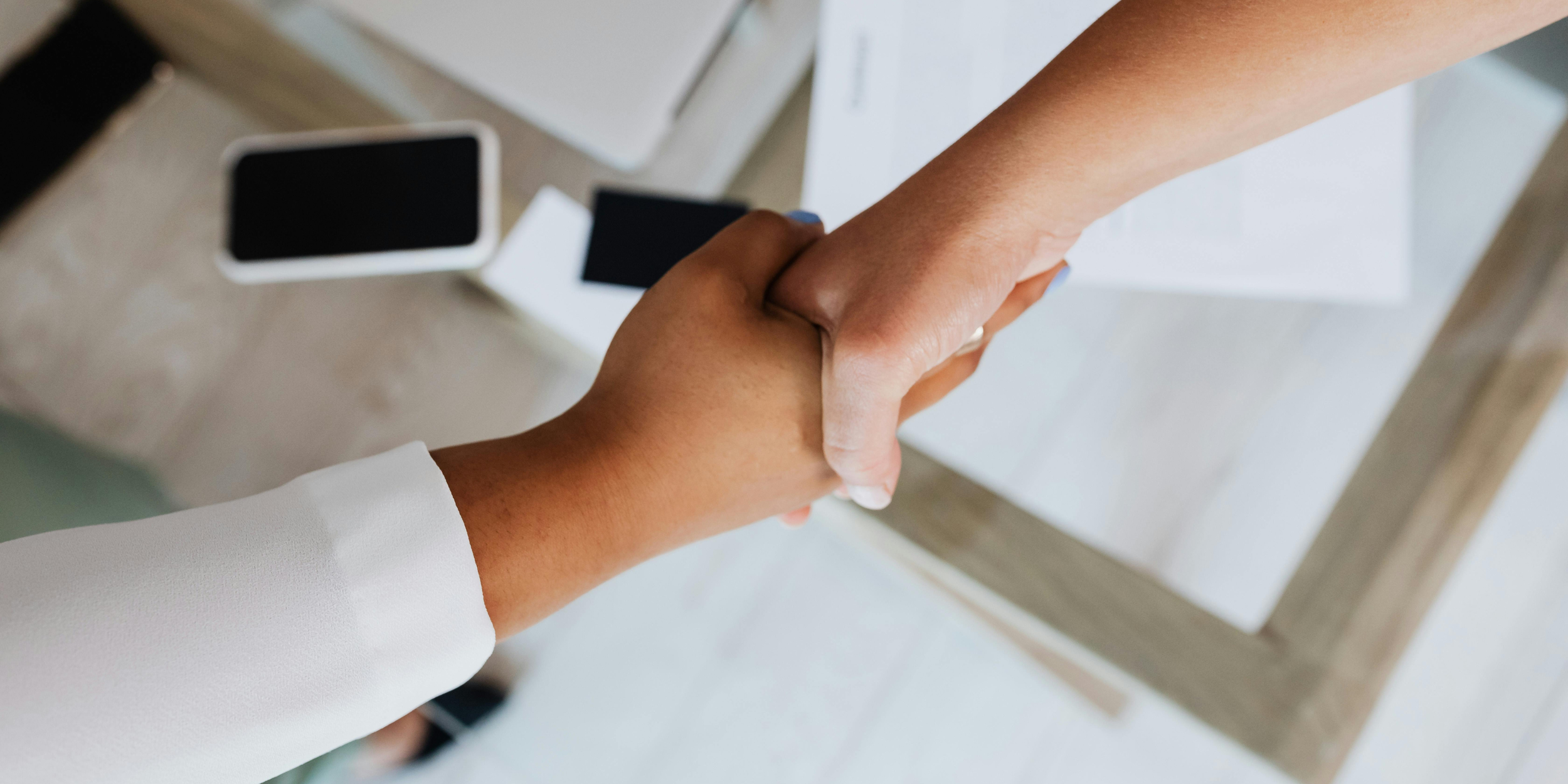 Two people shaking hands over a desk with a smartphone, notepad, and paper on it.