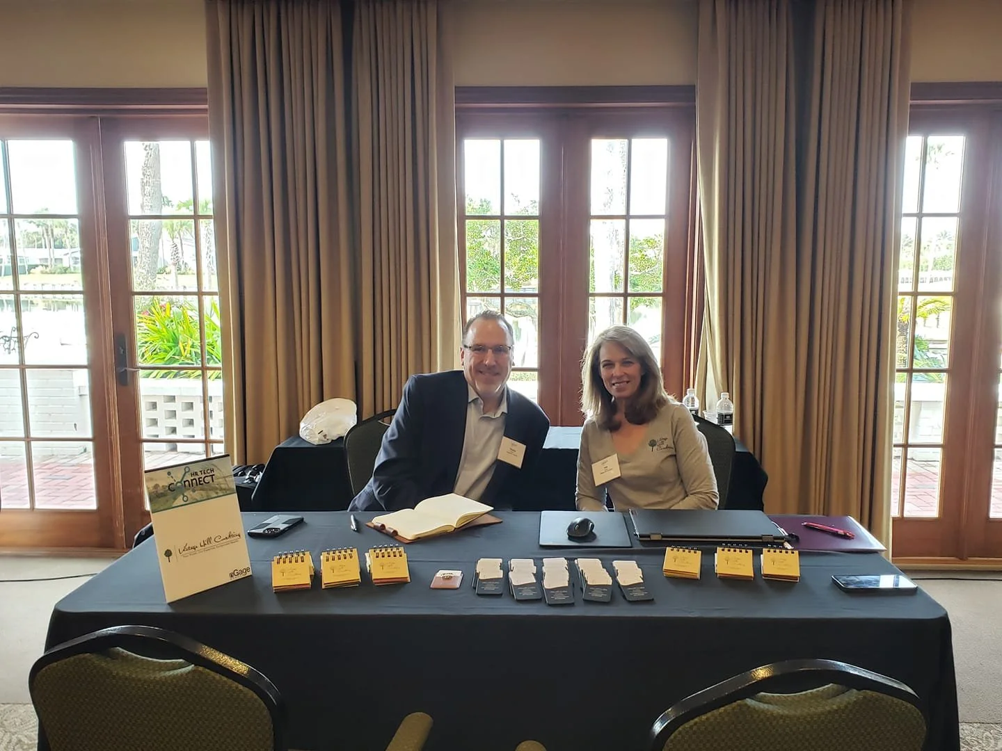A man and a woman sit at a table in front of large windows with beige curtains. The table has brochures, notebooks, smartphones, and water bottles. The man is wearing a dark suit and glasses, and the woman is wearing a light-colored top with a logo. They are smiling at the camera.