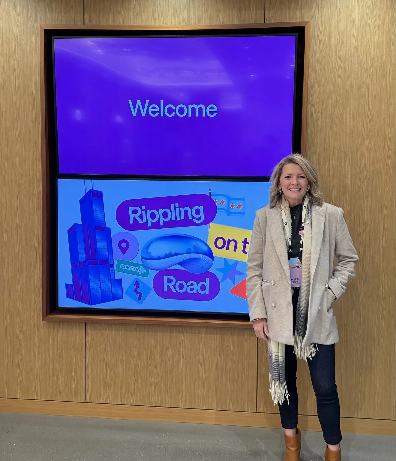 A woman standing in front of two digital screens, one displaying a welcoming message and the other showing colorful graphics related to urban environment, with the woman smiling and dressed in a light-colored coat and dark pants.