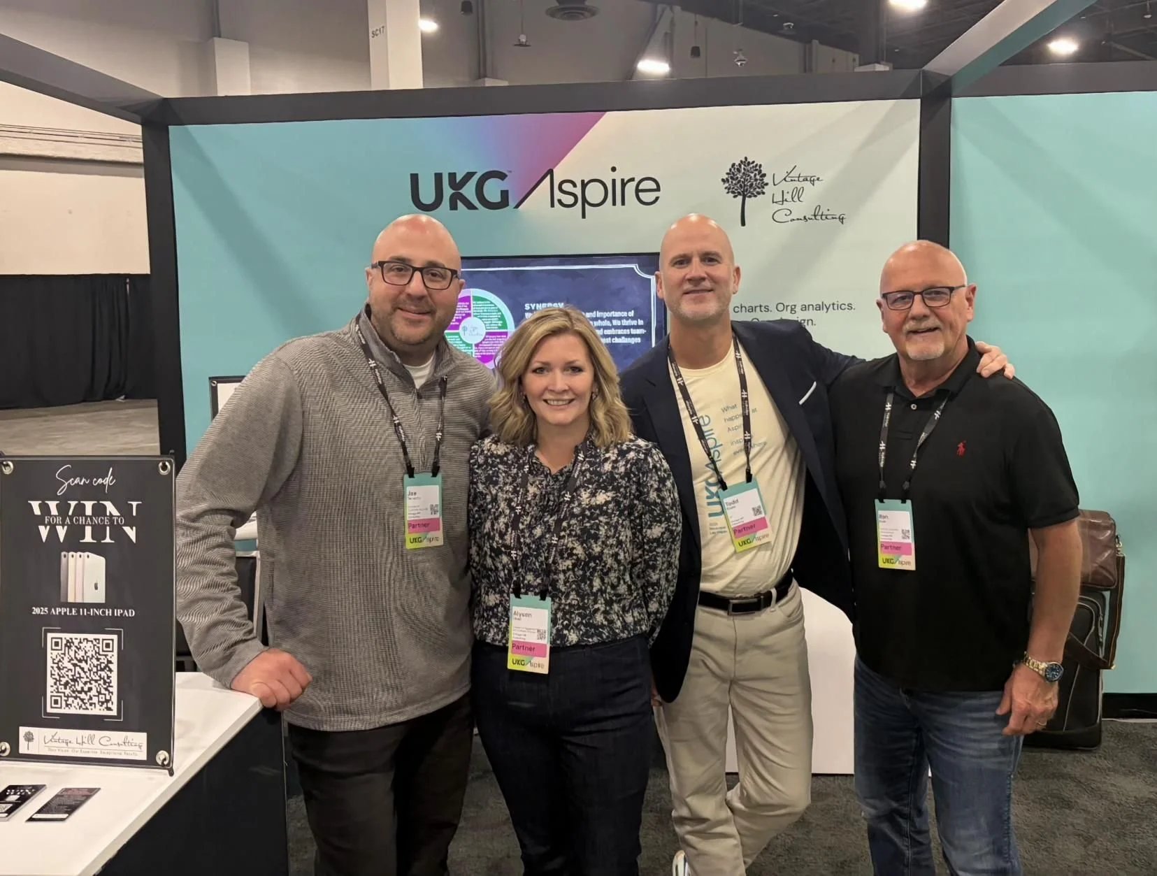 Four people standing together at an booth or conference, smiling at the camera. They are wearing conference badges. The backdrop features the text 'UKG Inspire' and branding. A QR code and promotional sign are visible to the left.