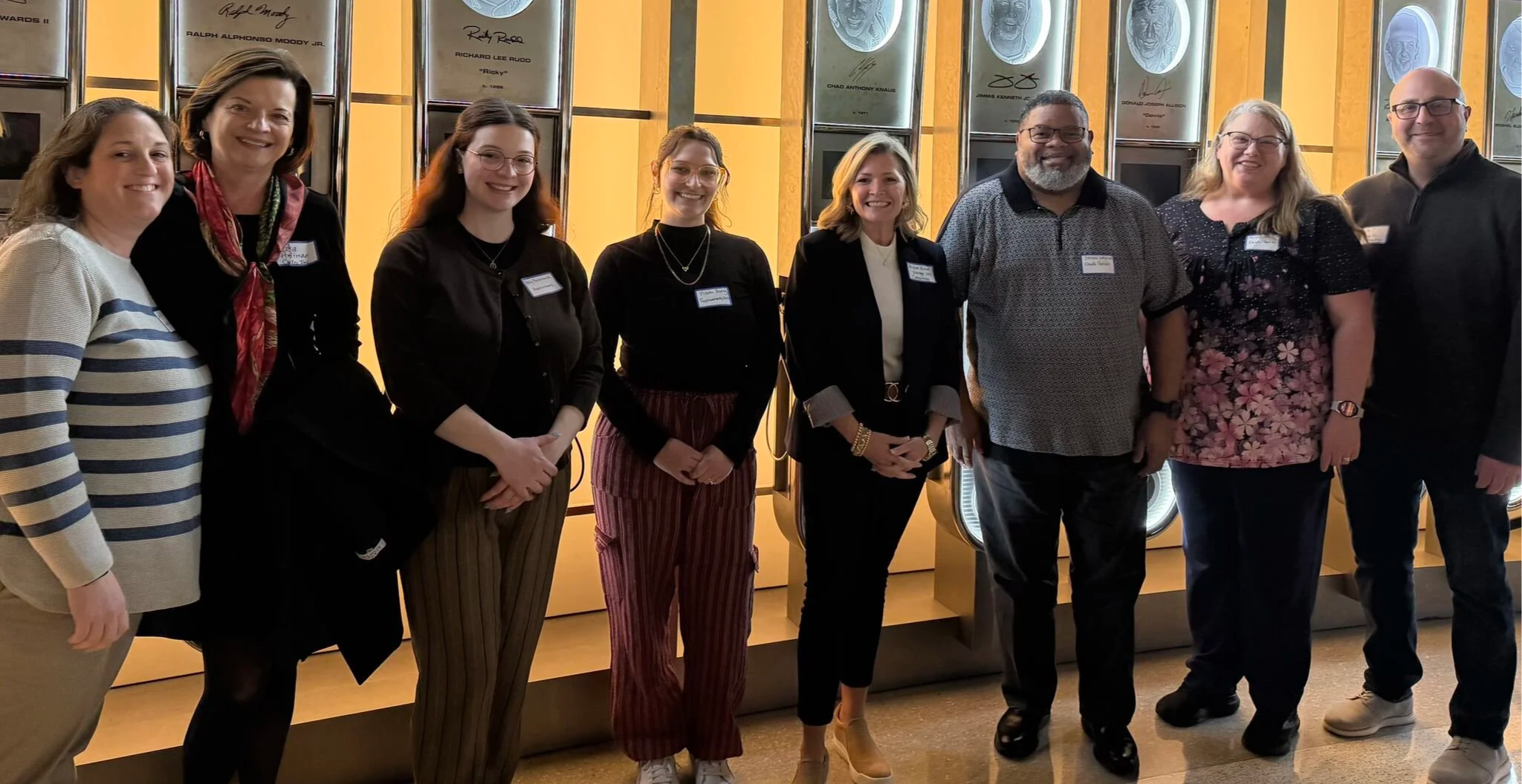 Group of nine people standing together inside a building with a wall of framed awards or signatures behind them. They are smiling and dressed in business casual attire.