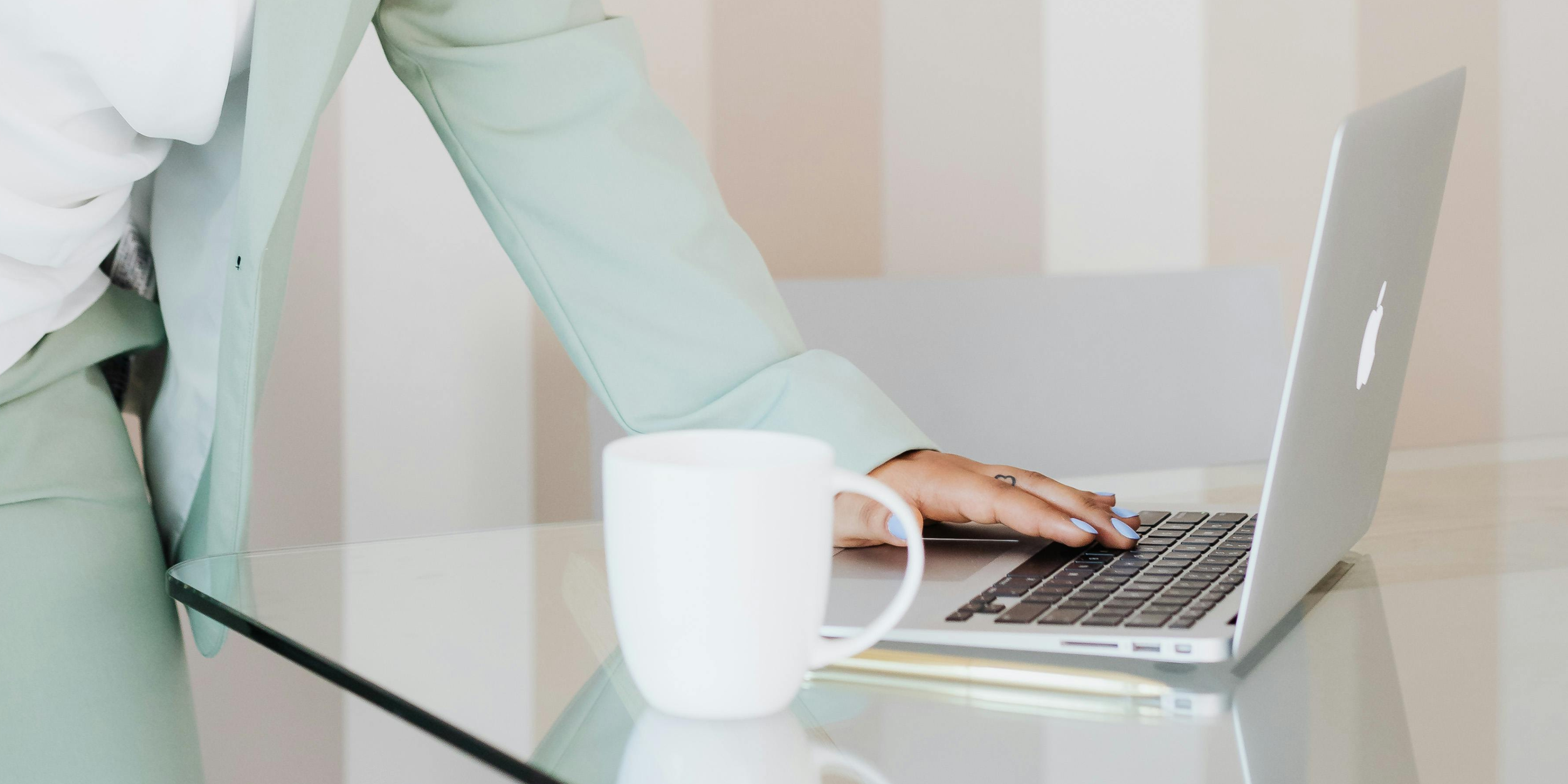 Person typing on a silver MacBook with a white coffee mug on a glass table in a bright room.