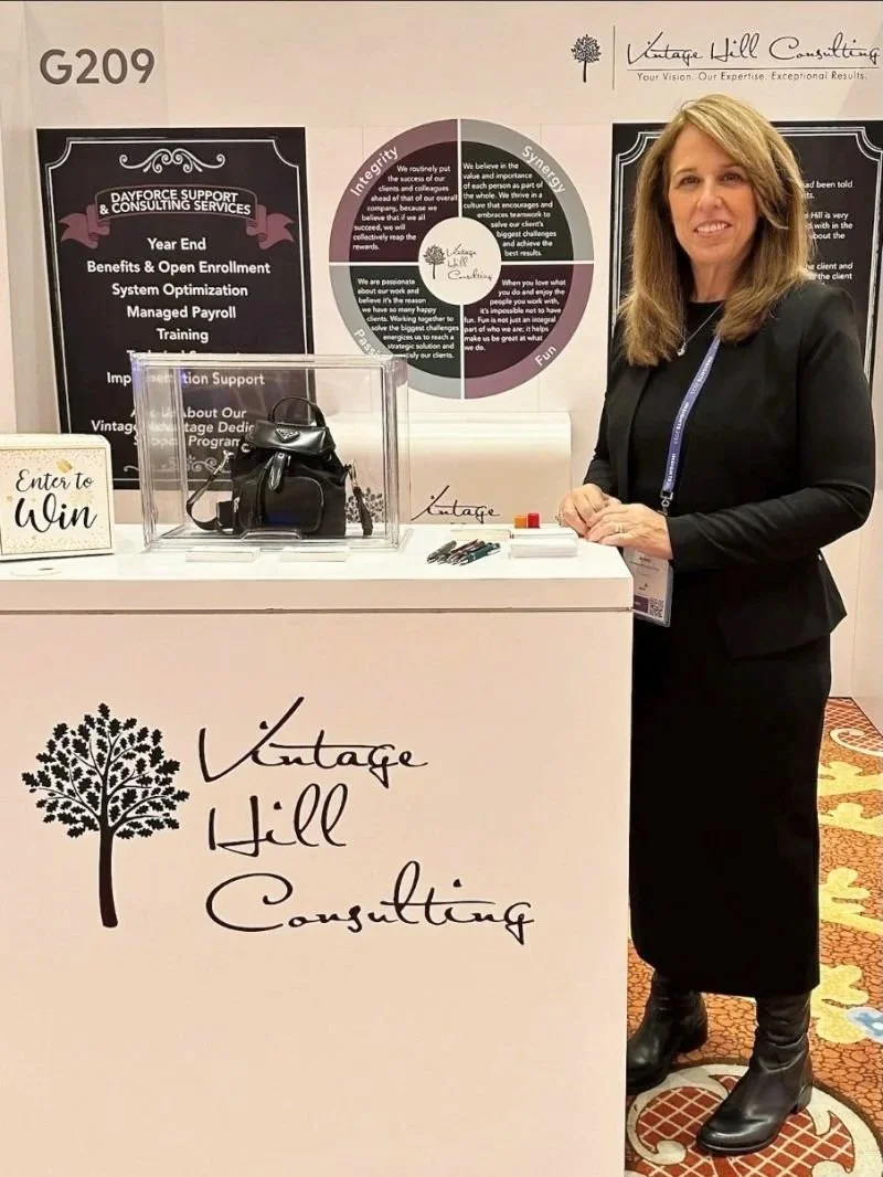 A woman standing behind a consulting booth for Vintage Hill Consulting, which features a large sign with a tree logo and the company name. The booth has a clear display case with a small black bag, a sign that says 'Enter to Win,' and some pens and markers. In the background, there are informational posters and banners about the company's services, including support, enrollment, payroll, and training.
