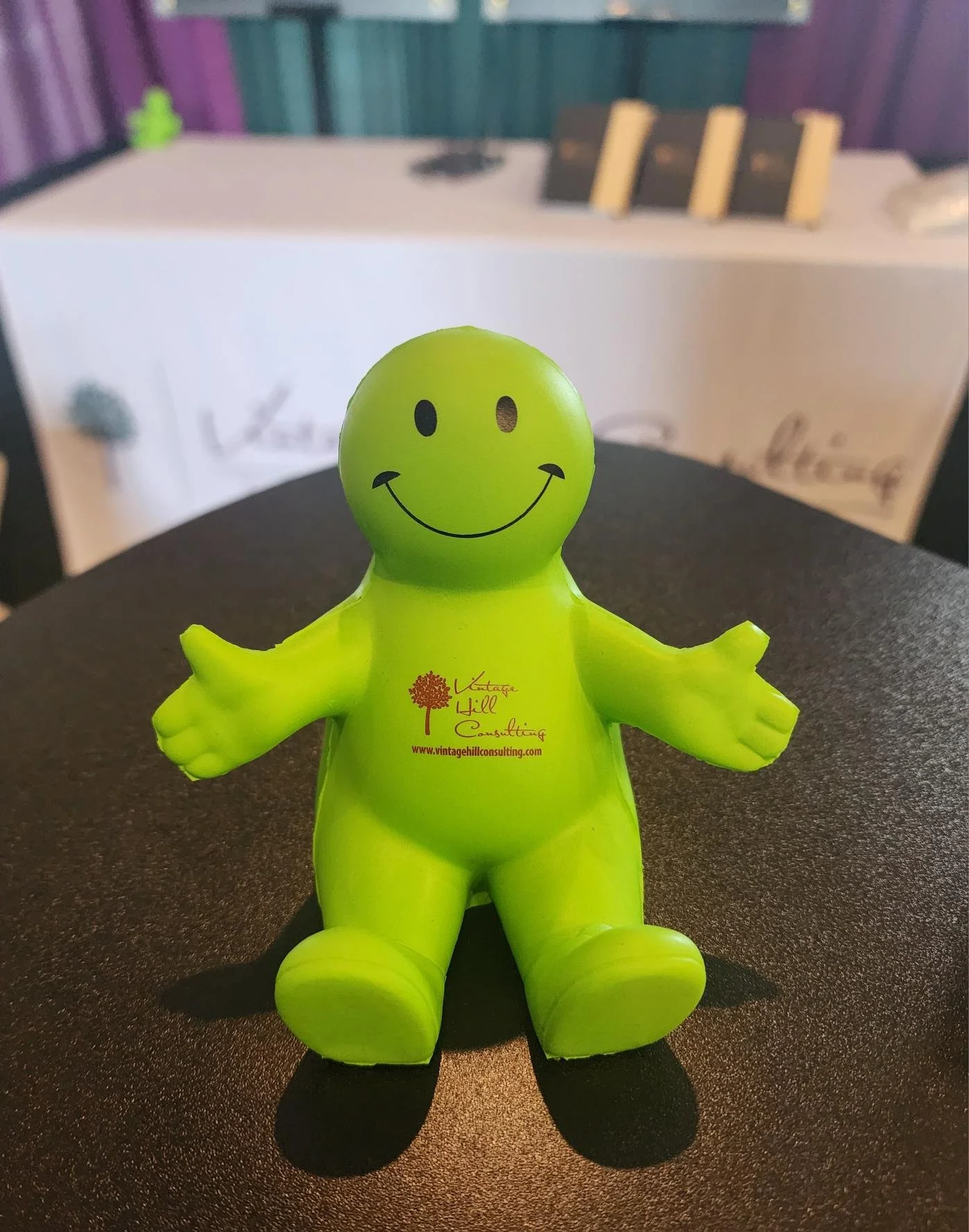 A green, smiling inflatable rubber toy with outstretched arms, sitting on a dark surface. The toy has a red logo and website printed on its chest. In the blurred background, there is a table with books and other objects.