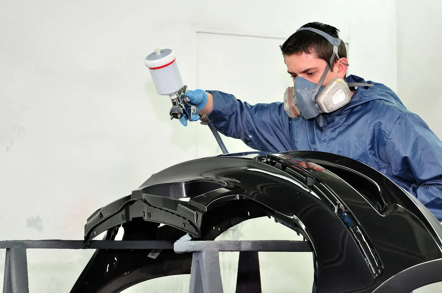 Person spray painting a black car fender, wearing protective mask, gloves, and blue coveralls.
