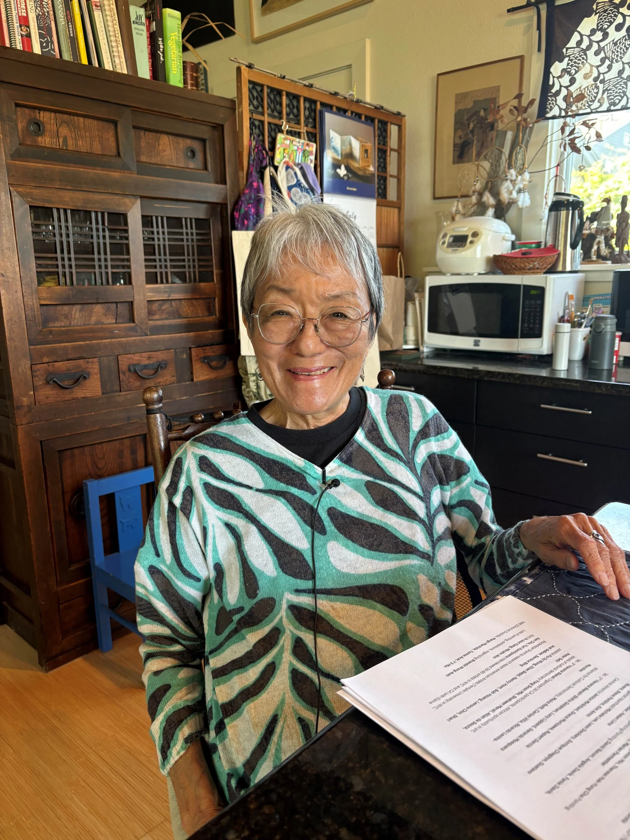 A female Japanese artist with short gray hair and glasses smiling at the camera while seated at a kitchen table, with a glass of water, papers, and a black electronic device in front of her. The kitchen has wooden furniture, a rice cooker and artwork