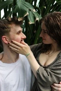 A young man and woman looking at each other with the woman touching the man's face, in front of leafy indoor plants.