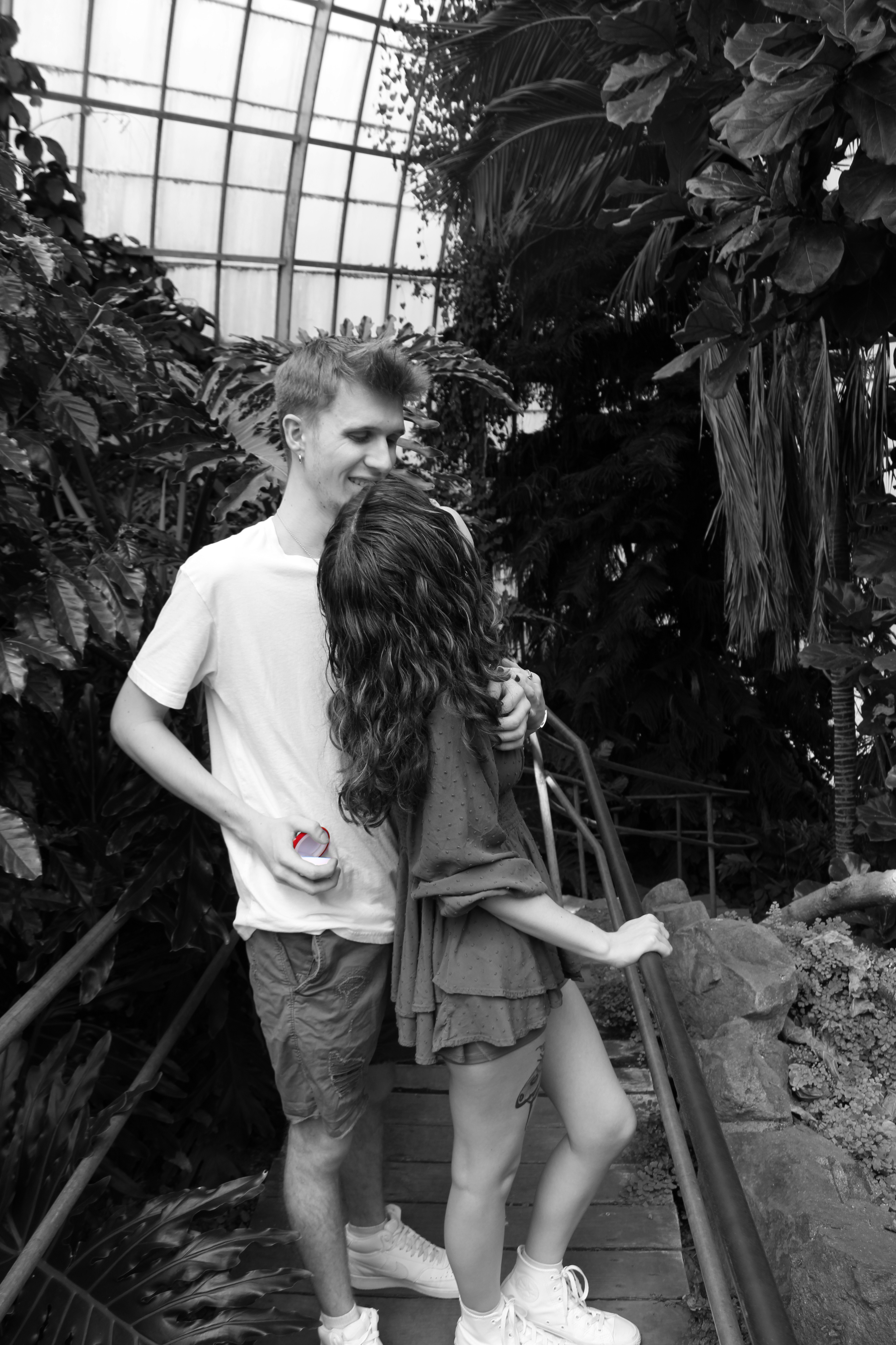 A man with short hair wearing a white t-shirt and shorts, smiling and holding a spray can, standing next to a mannequin of a woman in a dress inside a greenhouse filled with large tropical plants.