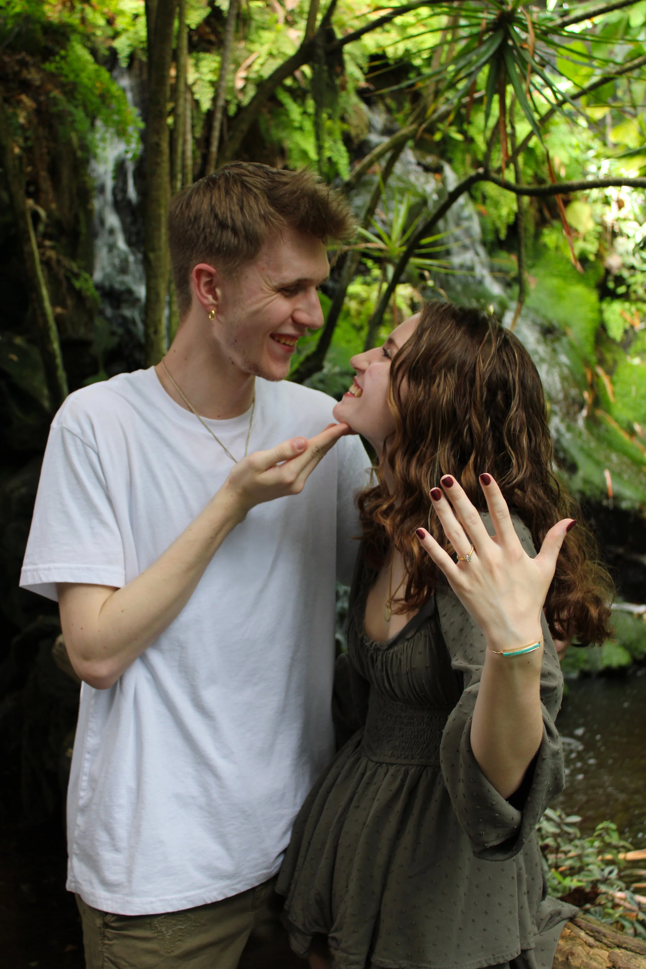 A young man and woman smiling at each other in a lush green forest, with waterfalls in the background.