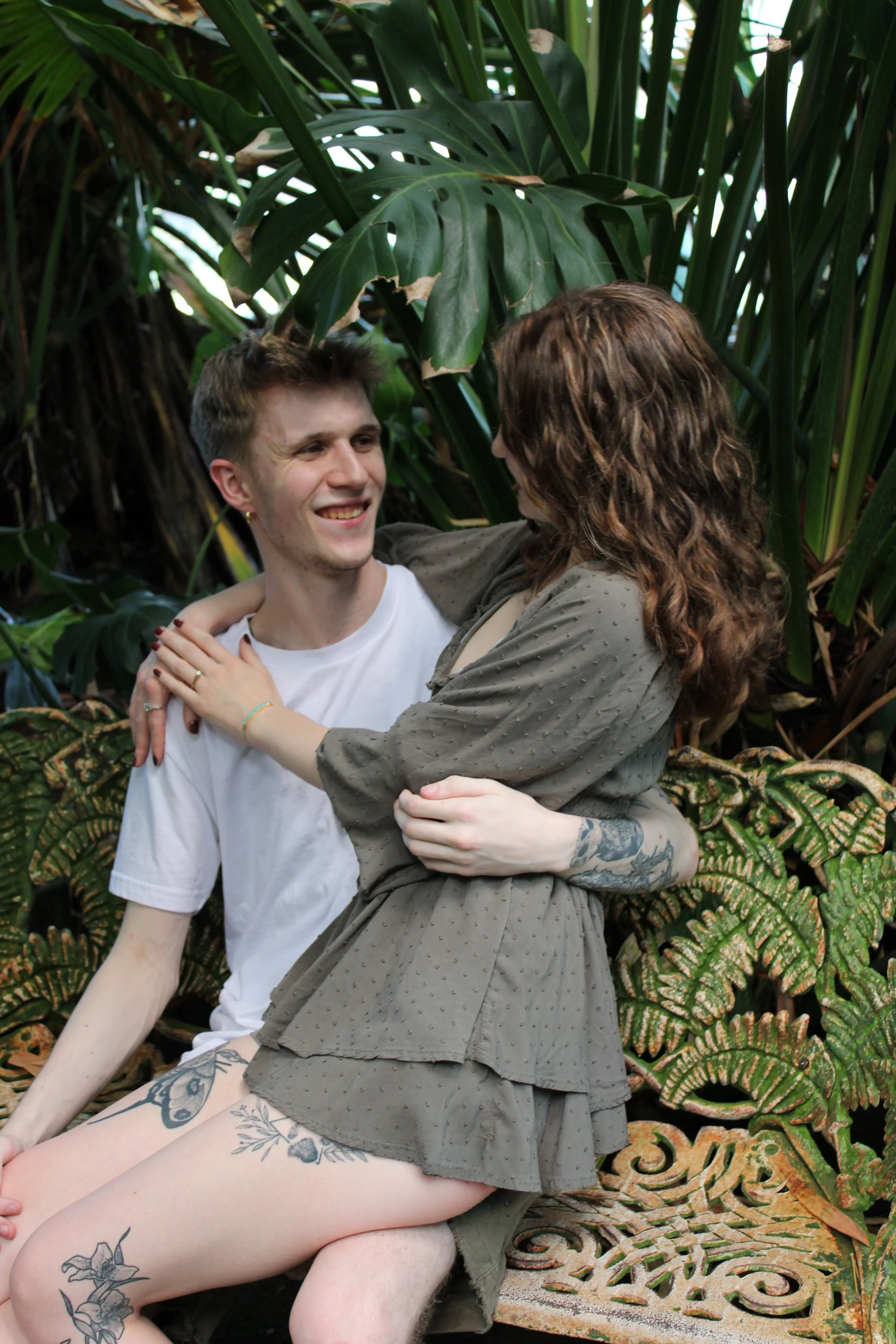 A couple sitting on a decorative garden bench surrounded by lush tropical plants, sharing a joyful moment and looking at each other.