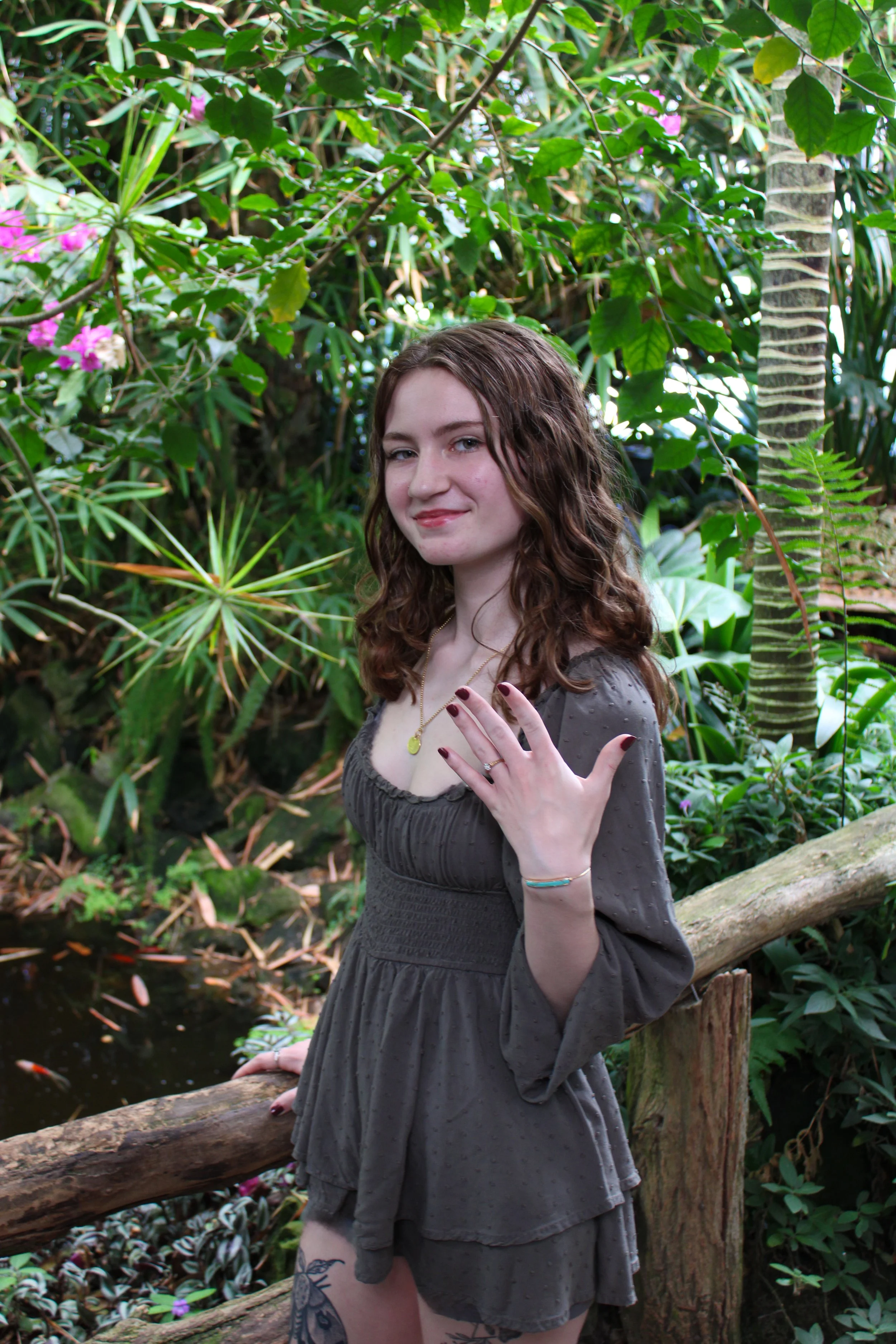 A woman with wavy brown hair wearing a gray dress standing in a lush green garden with pink flowers and various tropical plants, leaning on a wooden railing.