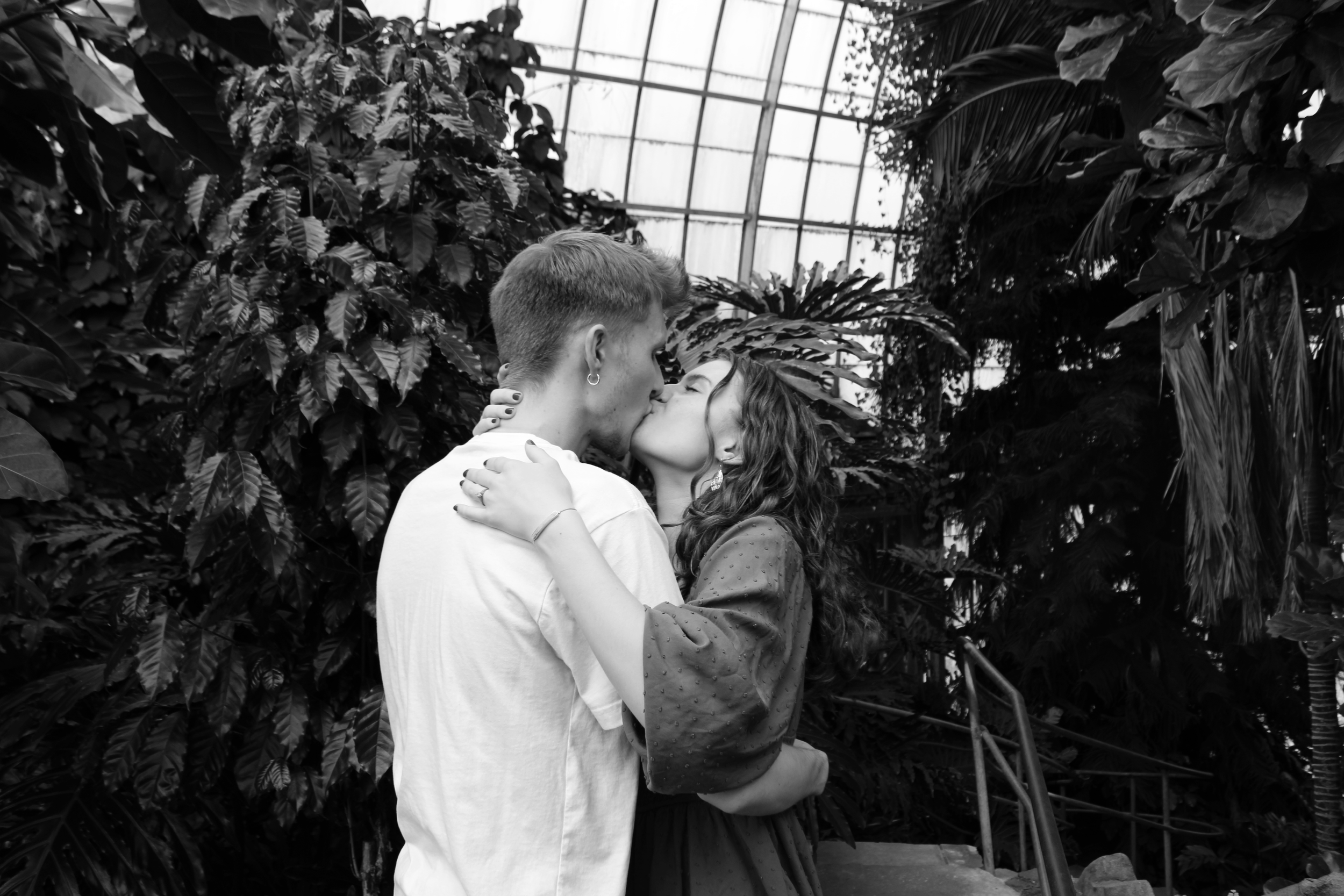 A young couple kissing in a greenhouse filled with large leafy plants.
