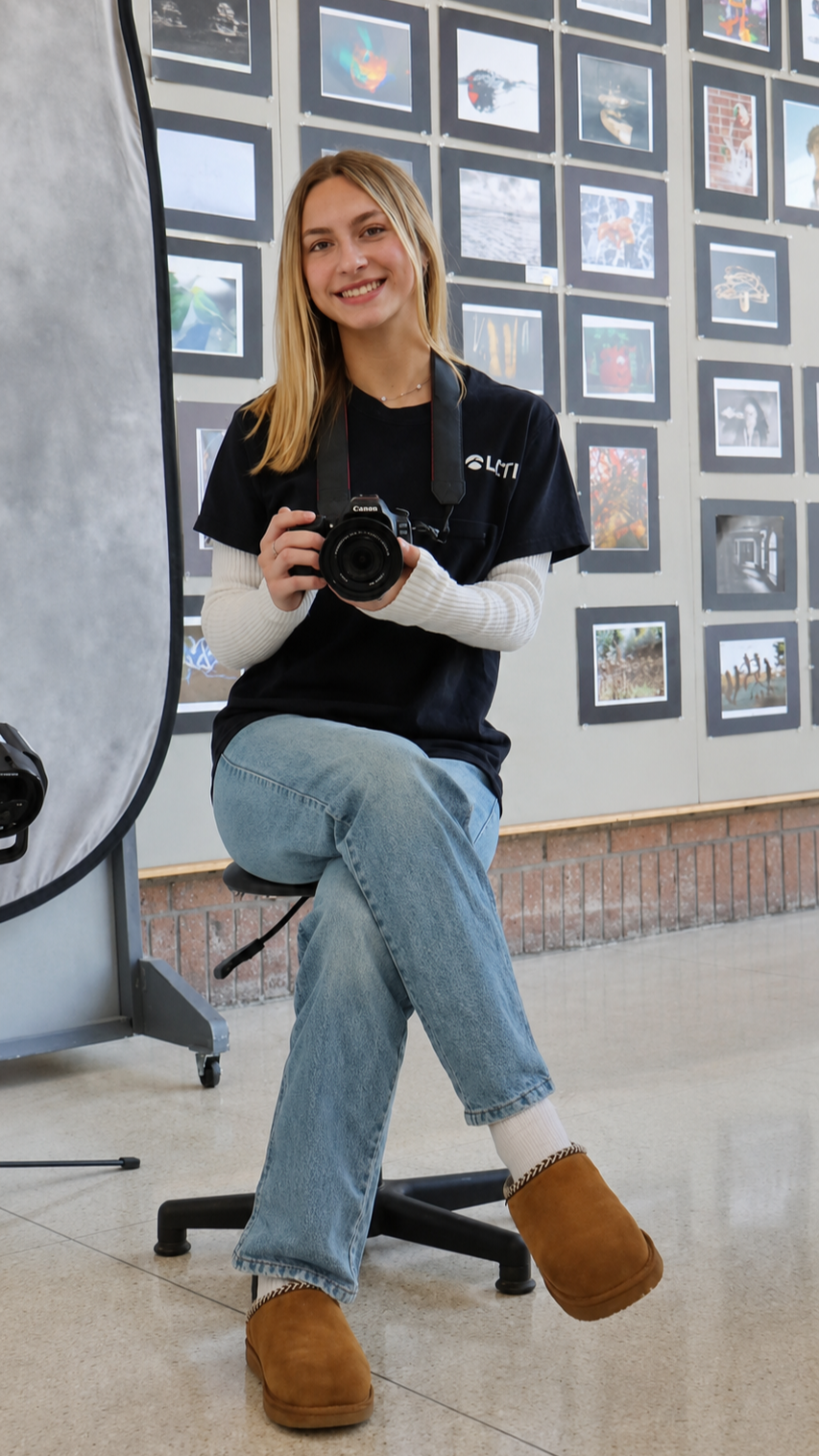 A young woman with blonde hair and a smile, holding a Canon camera, sitting on a stool in front of a gallery wall of framed photographs.