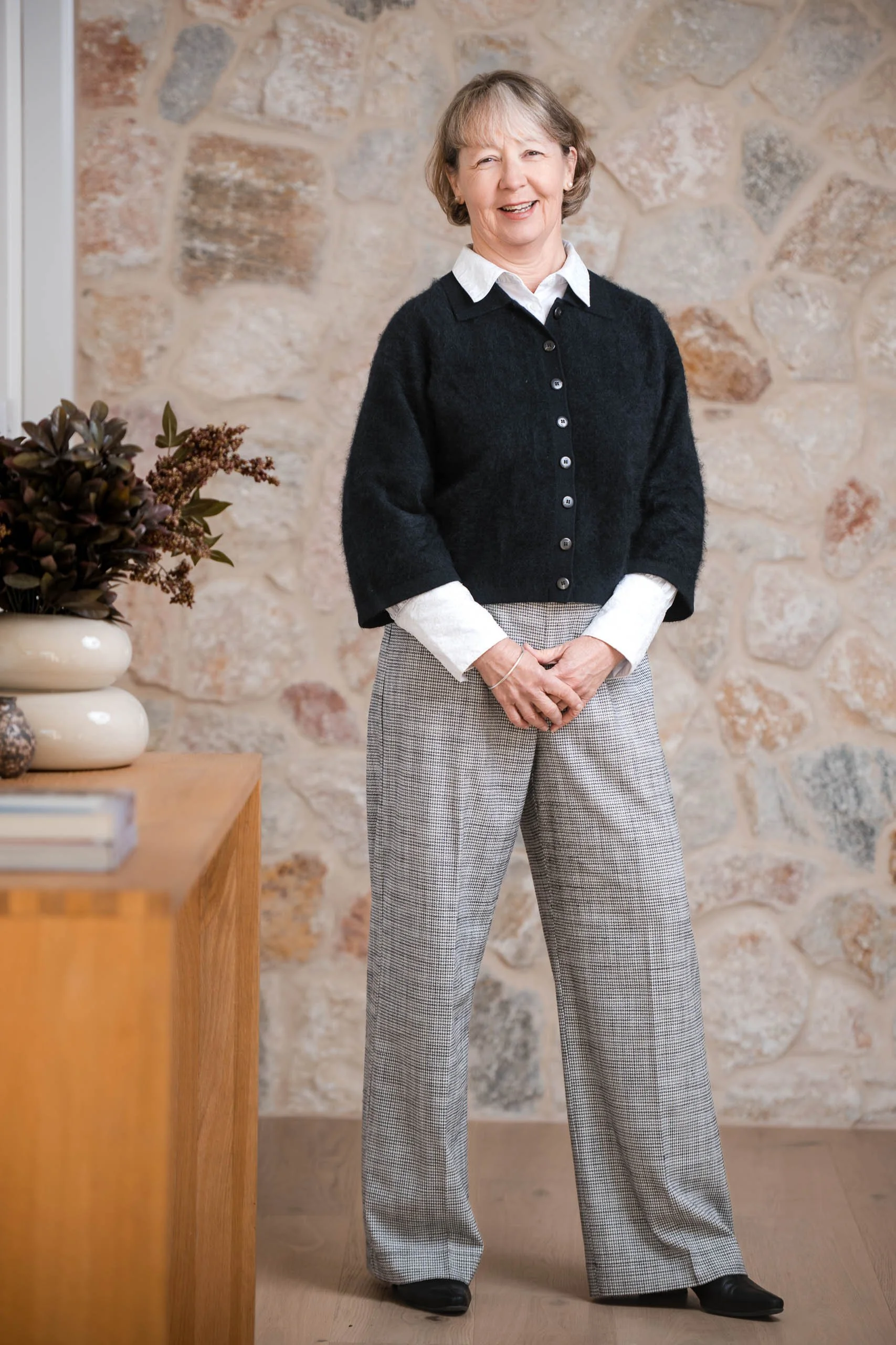 A woman standing indoors near a stone wall, smiling with her hands clasped in front of her. She is wearing a black cardigan over a white shirt, and gray checkered pants.