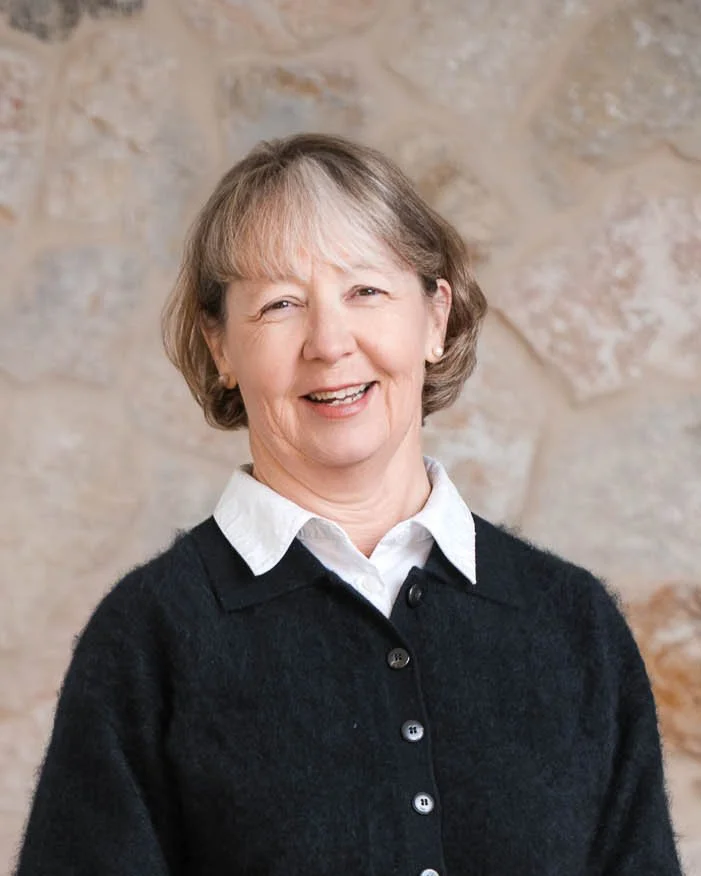 A smiling middle-aged woman with short, light brown hair, wearing a white collared shirt and a black cardigan, standing against a stone wall background.