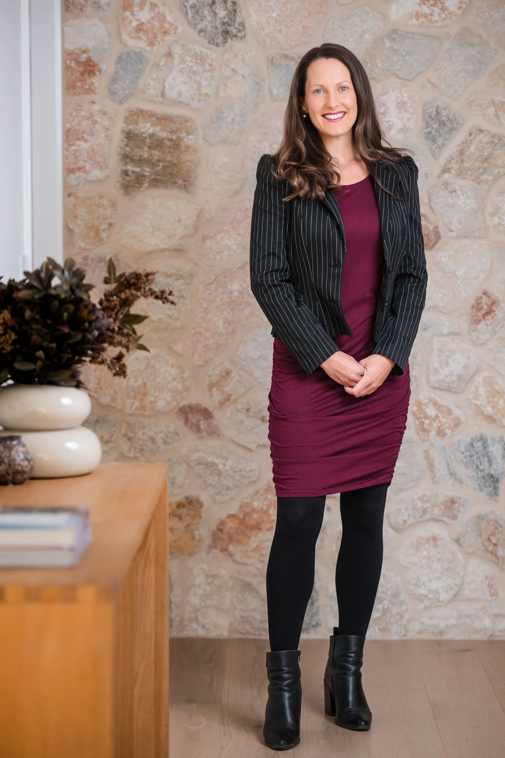 A woman with long brown hair, wearing a pinstripe blazer, maroon dress, black tights, and black boots, standing in front of a stone wall with a small table to her left holding decorative objects and books.
