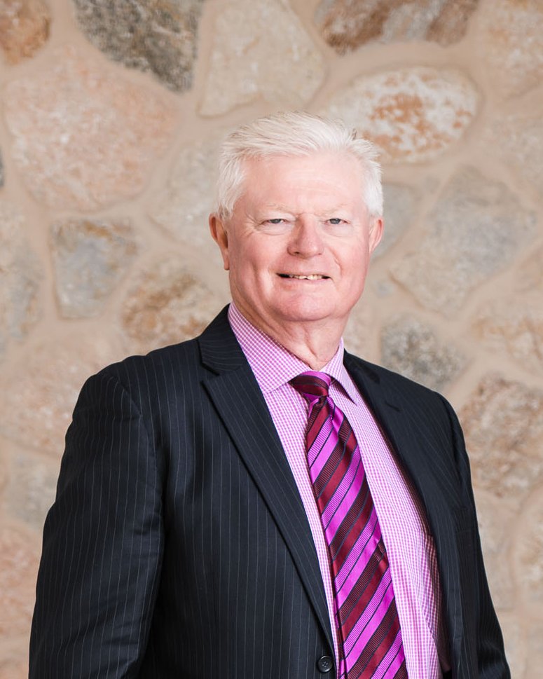 A man with white hair wearing a dark pinstripe suit, pink checkered shirt, and pink-striped tie, standing in front of a stone wall background.