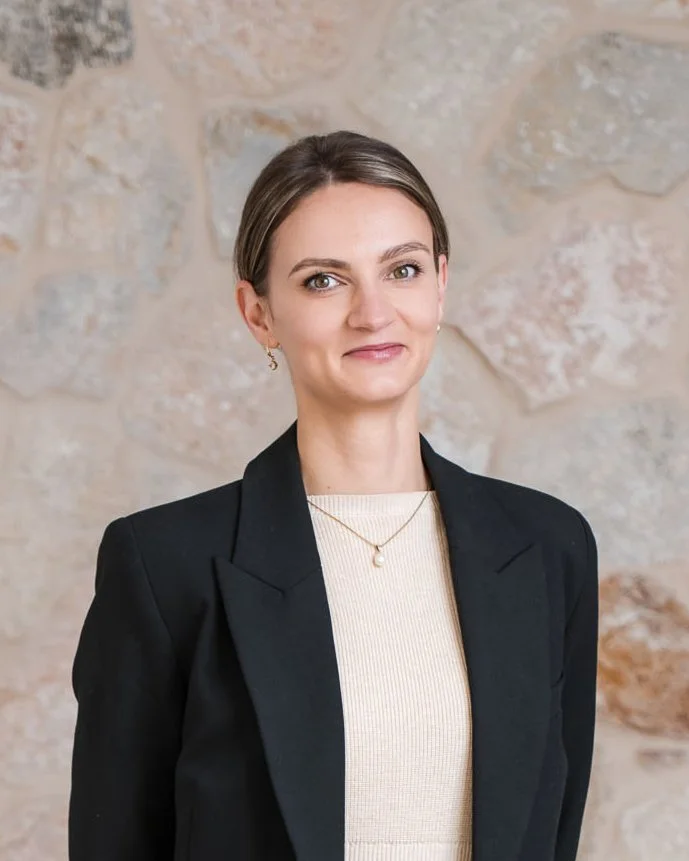 A woman with short brown hair and light skin standing in front of a stone wall, wearing a black blazer, a cream-colored top, and a gold necklace with a pearl pendant.