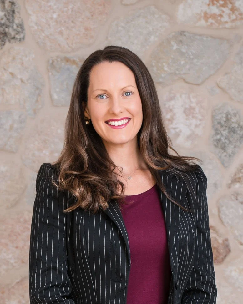 A woman with long brown hair, blue eyes, wearing a black pinstripe blazer over a burgundy top, standing in front of a stone wall, smiling.
