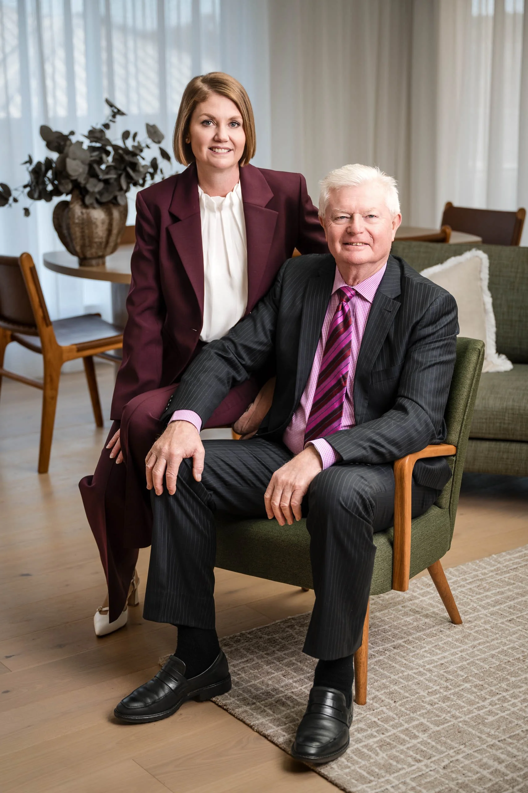 A middle-aged woman with short brown hair and a man with white hair posing together in a living room, both smiling at the camera. The woman is wearing a maroon blazer and white blouse, while the man is in a dark pinstripe suit with a pink shirt and striped tie. They are seated and standing near a chair and a side table with a plant in a vase.