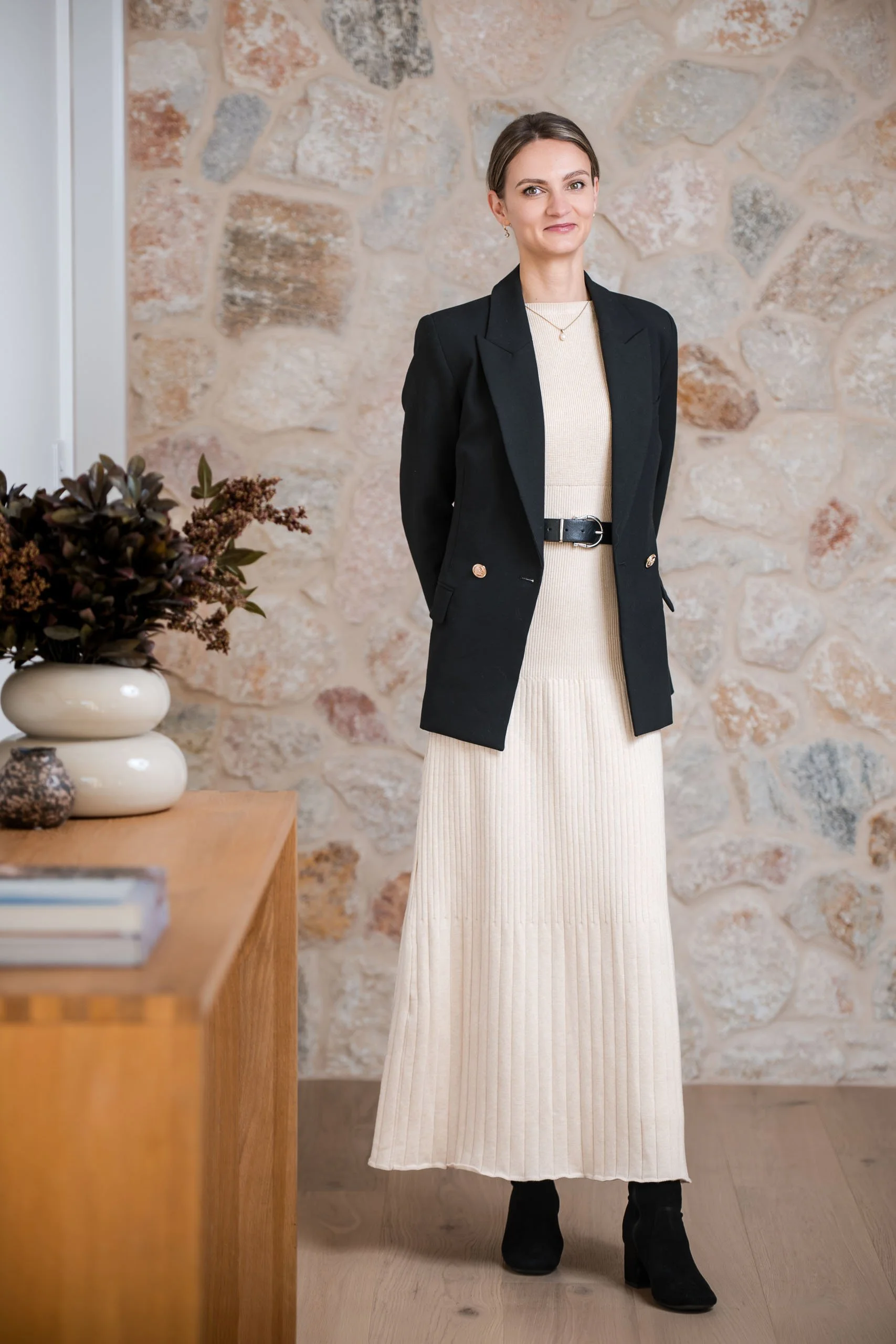 A woman stands indoors in front of a beige stone wall, wearing a black blazer over a cream-colored ribbed dress, with black boots. To her left, on a wooden table, there is a white vase with dried dark purple flowers and a stack of books.