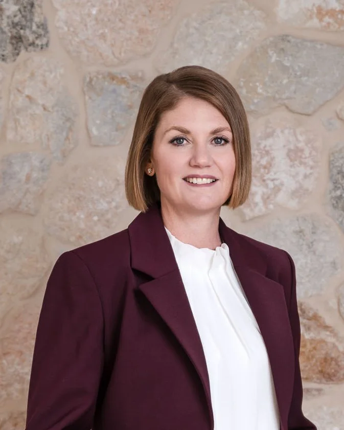 A woman with shoulder-length brown hair wearing a white blouse and a dark burgundy blazer, standing in front of a stone wall.