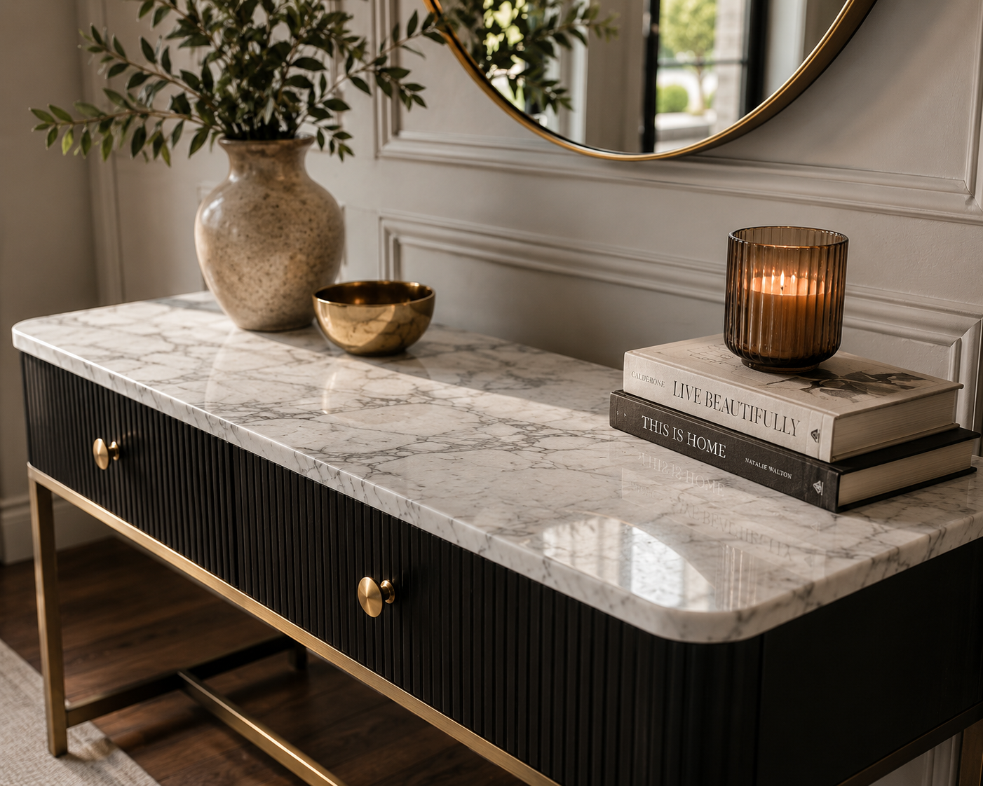 A modern black sideboard with gold accents has a white marble top. On the top, there is a large beige ceramic vase with green leaves, a small gold bowl, a lit amber candle in a ribbed glass holder, and two stacked books titled "Live Beautifully" and "This Is Home".