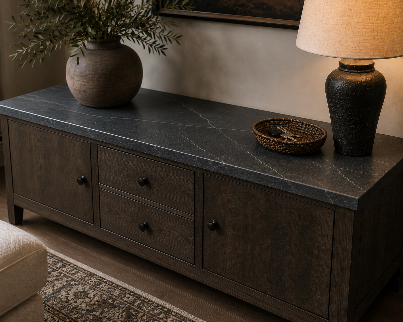 A dark wooden sideboard with a black marble top, decorated with a large brown vase with greenery, a round basket with keys and a keychain, and a black ceramic table lamp with a beige lampshade.