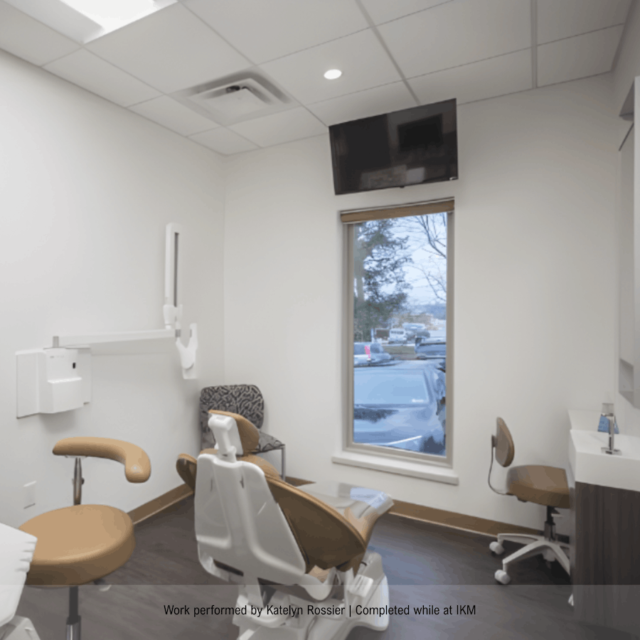 Medical examination room with a dental chair, a chair in the corner, a window showing trees and parked cars, a wall-mounted TV, and a sink with a chair nearby.