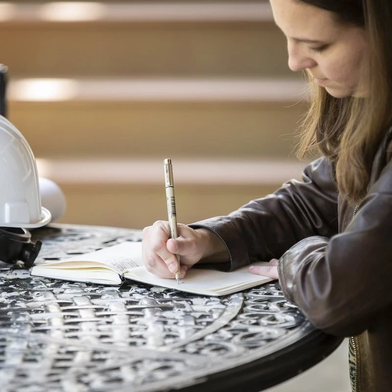 Katelyn taking notes in a book sat at a table with a hard hat.