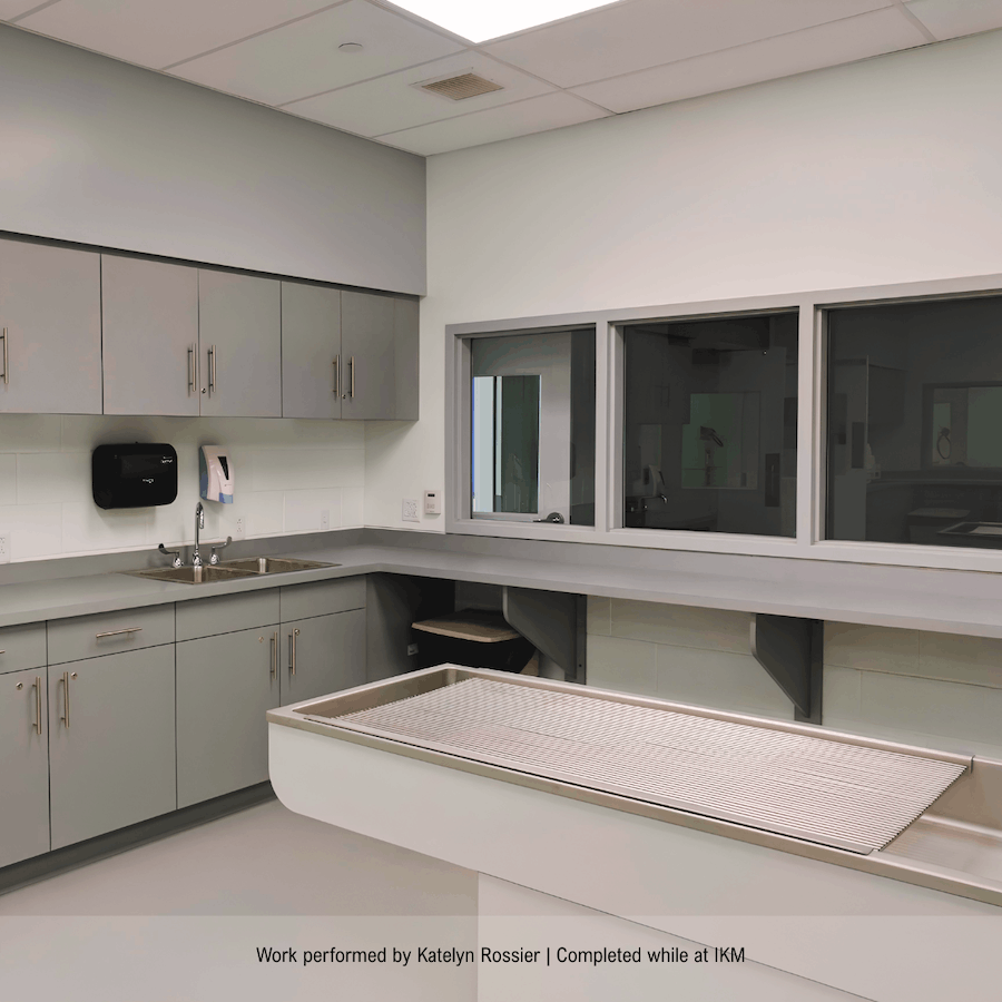 A sterile hospital room with gray cabinetry, a sink, a soap dispenser, a window, and a padded examination table.