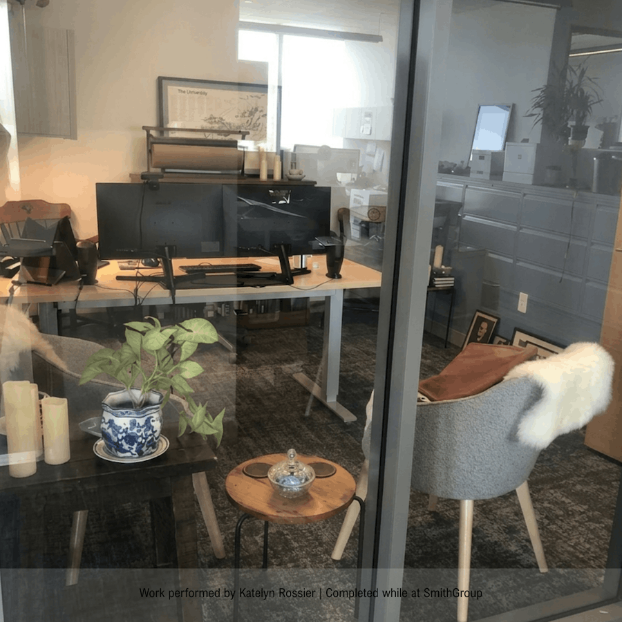 Office workspace seen through glass wall, with computer monitors, a desk, chairs, and decorative items including candles, framed photos, and plants.