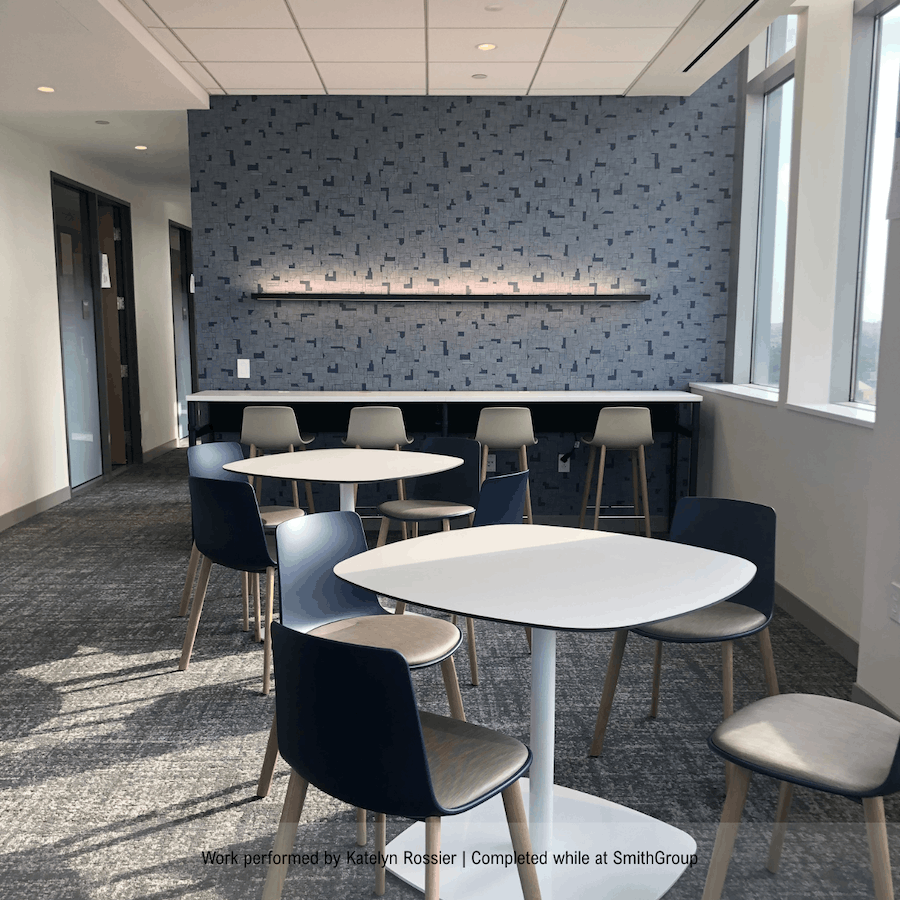 Modern office lounge or break room with round tables, black and beige chairs, a blue textured accent wall, and large windows letting in natural light.