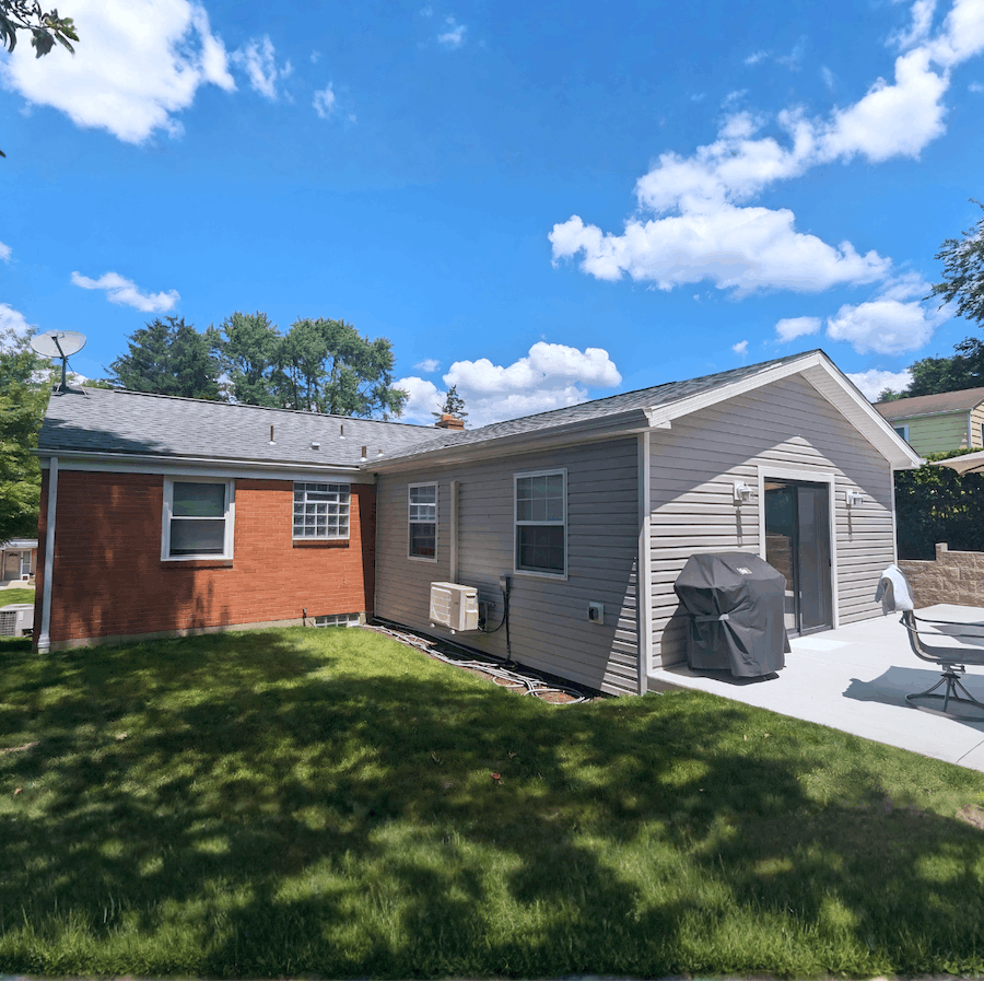 Backyard of a house with green grass, a concrete patio, patio furniture, a grill, and an air conditioning unit, with a blue sky and scattered clouds overhead.