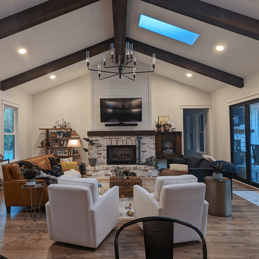 A living room with a vaulted ceiling, wooden beams, and a skylight. There is a brick fireplace with a flat-screen TV mounted above it. The room has two white armchairs facing the fireplace, a tan leather sofa, and a dark sofa. A chandelier hangs from