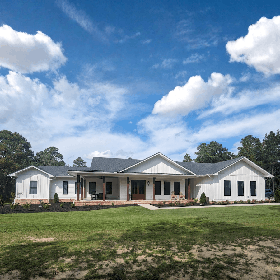 Modern white house with a gray roof, large front porch, and landscaped yard under a blue sky with clouds.