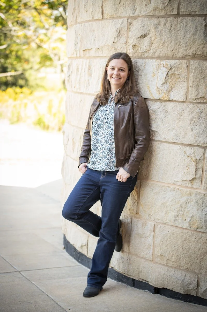 Katelyn in a leather jacket and jeans leaning against a wall outside