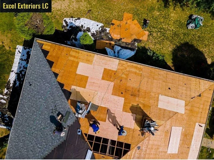Aerial view of a house under roof renovation, with workers installing new plywood roofing. The house has a gray shingled section and a large area of exposed plywood, with construction materials and tools nearby.